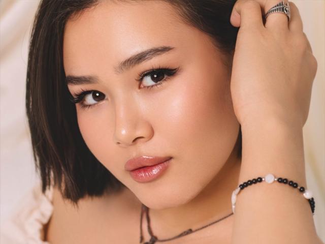 Close-up of ArianaFlower with medium skin tone, dark brown shoulder-length hair, and glossy pink lips. She has black eyeliner, slightly arched eyebrows, and a subtle, shiny complexion. Her right hand is raised, touching her hair, showing a black beaded bracelet and a silver ring. Soft, warm light highlights her facial features. She has a slightly serious, yet approachable expression. The image focuses on her face and upper shoulders.