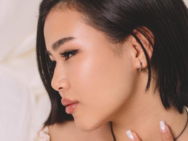 ArianaFlower's face, showing her smooth, tan skin, black bob, and subtle makeup, with a small nose ring and glossy pink lips. Her hand with white nails rests on her neck.