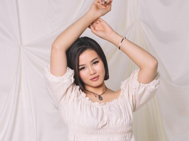 ArianaFlower has a medium skin tone and short black hair. She's wearing a light pink, puffed-sleeve, off-the-shoulder dress with a ruffled neckline. Her arms are raised, hands touching above her head. She has a small black pendant necklace and a thin bracelet on her right wrist. Her makeup includes dark eyeliner and pink lipstick. The soft white background has gentle folds. The image has a clean, professional look with a focus on ArianaFlower's relaxed, confident pose.