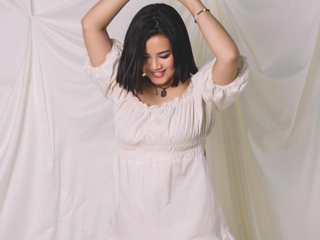 ArianaFlower with black bob, arms raised, smiling, wearing a white, short-sleeved, ruffled dress, necklace, and bracelet, standing against a white draped background.