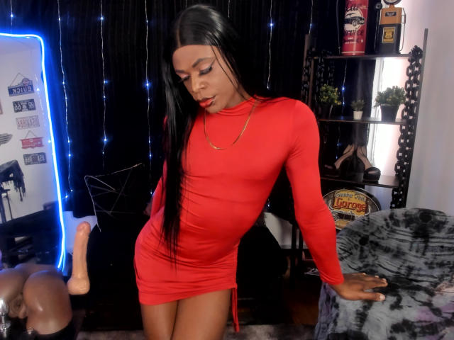 Venus Miller, avec de longs cheveux noirs, portant une robe rouge moulante, debout, penchée en arrière, montrant ses fesses, avec un sextoy à gauche et des étagères en arrière-plan..