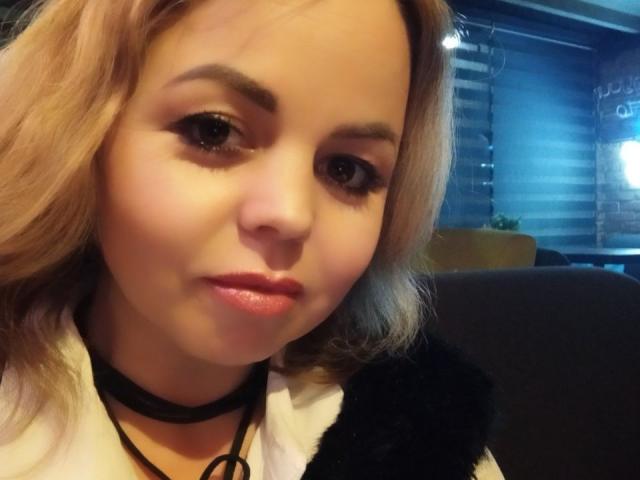 EvaMinni has blonde hair, brown eyes, and pink lipstick. She's wearing a black choker and white shirt with a black furry shoulder. Her face is slightly flushed.