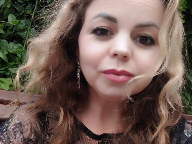 EvaMinni's face with wavy blonde hair, pink lipstick, and black lace top. She has brown eyes, subtle makeup, and is wearing dangling earrings. Her expression is neutral.