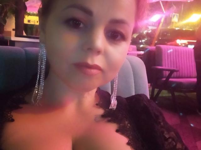 EvaMinni with pinkish-purple lighting, wearing black lace and diamond earrings, showing her tits in a low-light setting, looking directly at the camera with a neutral expression.