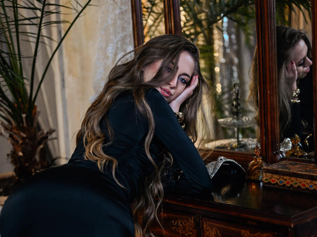 GoGoDream with long brown hair, black dress, and butt leaning on a wooden mirror, looking seductively over her shoulder, hand on cheek, in a dimly lit room.