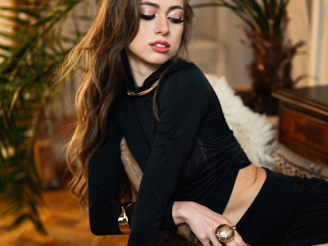 GoGoDream leans forward on a dark, wooden surface, eyes closed, lips slightly parted. She has long, wavy brown hair, wearing a black long-sleeve crop top and black pants. Her right hand, adorned with a gold bracelet and ring, rests on the surface. She has fair skin, pink lipstick, and subtle, metallic eyeshadow. The background includes blurred green plants and a white, fluffy pillow. The image uses warm, natural lighting. The composition emphasizes GoGoDream's relaxed, sensual posture.