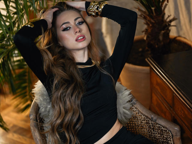 GoGoDream mit langen braunen Haaren, schwarzem Oberteil und goldenen Armreifen, Hände im Haar, blickt in die Kamera, sitzt auf einem Korbstuhl, sexy, süß, kleiner Nasenring, roter Lippenstift..