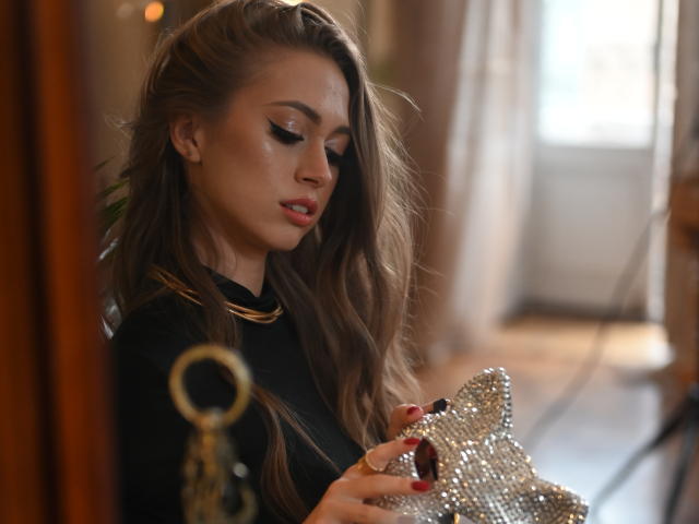 GoGoDream, a light-skinned woman with long, wavy brown hair, holds a silver, sequined clutch in her right hand. She wears a black top with a gold necklace. Her eyes are closed, lips slightly parted with pink lipstick. She has red nail polish and subtle, shimmery eyeshadow. The image has a shallow depth of field with a blurred background. Light filters through a window behind her. She has a focused, gentle expression. The photograph uses warm, soft lighting.