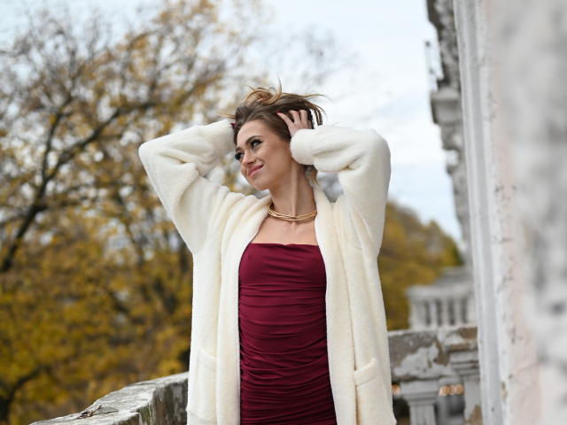 GoGoDream with hands in brown hair, wearing a white fur coat over a ruched red dress, smiling, standing on a stone balcony, autumn trees in the background.
