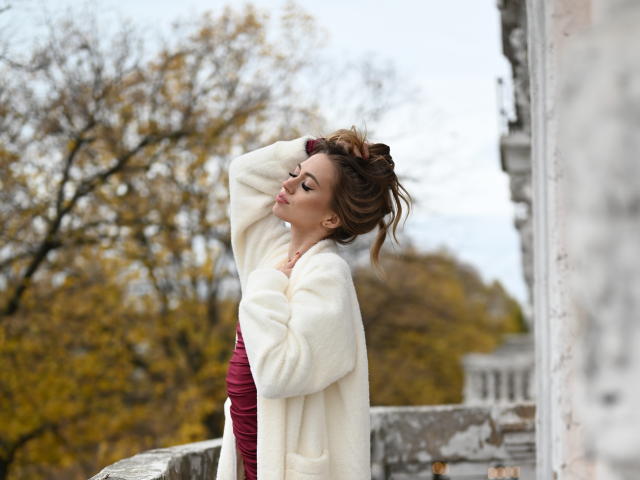 GoGoDream stands on a stone balcony, arms raised, hands in her brown, wavy hair. She wears a white, fluffy coat over a maroon dress. Her eyes are closed, lips slightly parted. Background shows blurry autumn trees. The right side has a weathered stone wall. The image has a soft, natural light. GoGoDream's relaxed, confident posture contrasts with the textured, aged stone. The color palette is warm and muted.