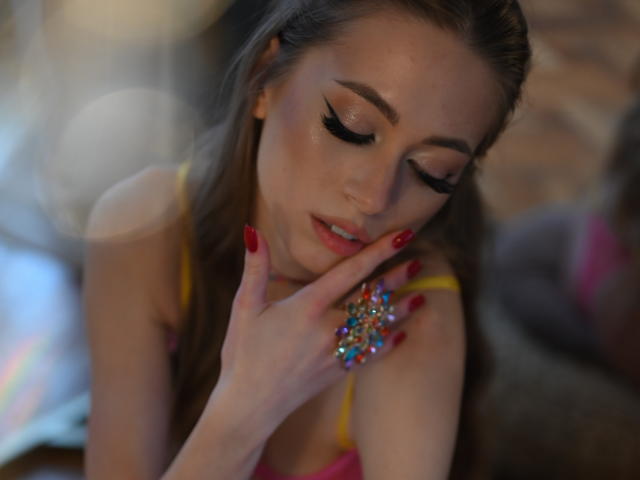 GoGoDream with her eyes closed, touching her lips with a hand wearing a colorful ring, has red nails and a pink top, looking sexy and seductive.