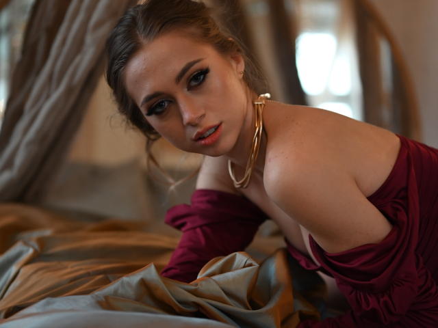 GoGoDream, con i capelli castani, il trucco scuro sugli occhi e gli orecchini dorati, indossa un abito rosso con una spalla scoperta, è seduta sul letto, inclinata in avanti, con lenzuola dorate e marroni. Il seno è parzialmente visibile..