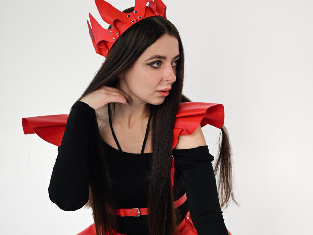 DiamondEyesss with long brown hair, wearing a red crown, black off-shoulder top, and red shoulder armor, looking to the side with a slight pout, hand near her face.