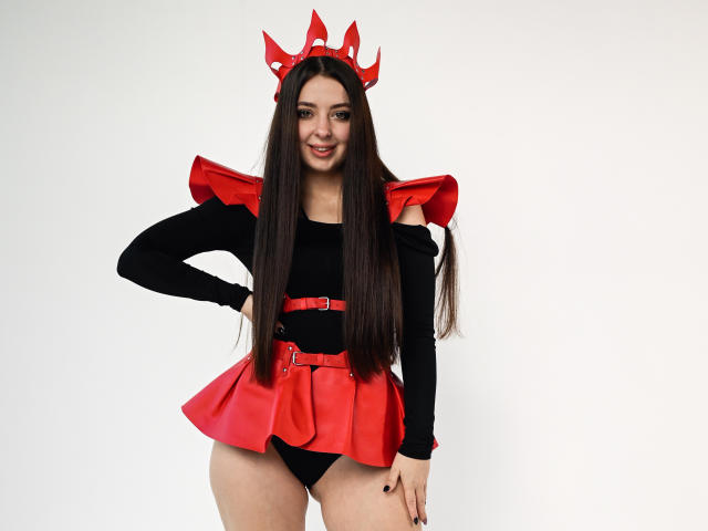 DiamondEyesss stands in a white background, wearing a red devilish headpiece with horns, a black long-sleeve top, and a red pleated skirt with a matching belt. She has long, straight black hair, dark eyebrows, and a slight smile. Her right hand rests on her hip, and she has black nails. The red and black colors contrast sharply, with the red horns and skirt drawing attention. Her overall look is playful and slightly provocative.