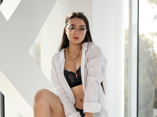 DiamondEyesss is a young woman with long dark brown hair, wearing clear glasses, an open white shirt, and a black lace bra. She has a gold chain necklace and is seated with one knee up. Her expression is neutral, and she's looking slightly to the right. The image is brightly lit with natural light coming from the right side. Her skin is light, and she has a slim build. The background includes a large white geometric shape on the left.