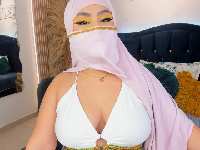 DaliilaAli with pink hijab, yellow eye makeup, white halter top showing big tits, gold necklace, and matching pink mask, sitting on dark blue tufted bed.