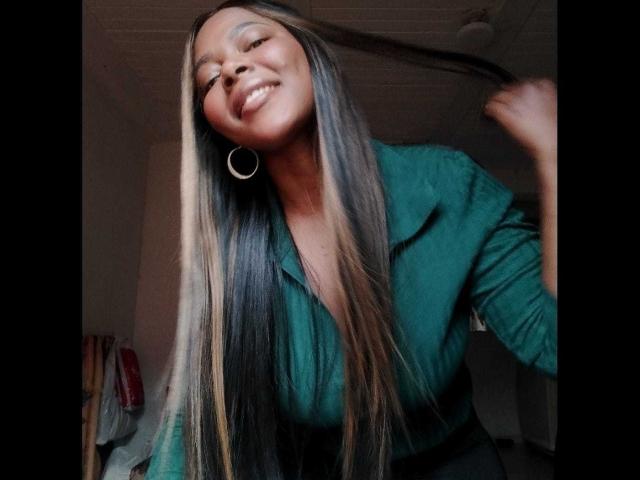 MacarenaBoobs with long straight blonde and black hair, green shirt, big hoop earrings, smiling, holding hair, dark skin, slight tilt, casual indoor setting.