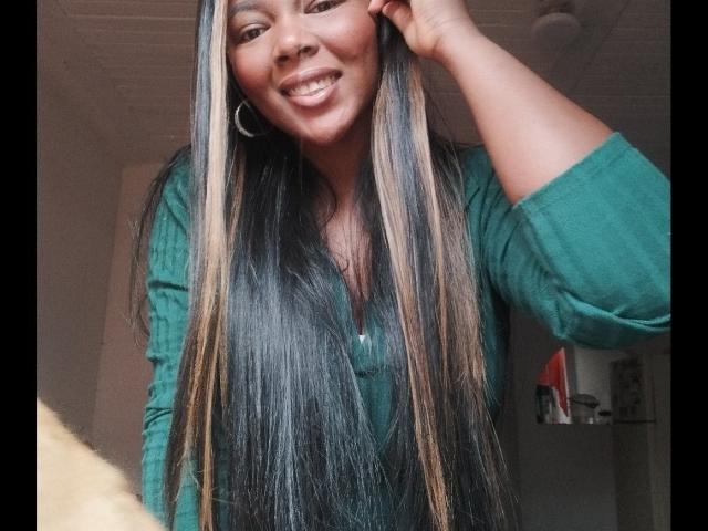 Macarena, avec de longs cheveux noirs et blonds, souriante, portant un t-shirt vert côtelé, touchant son oreille avec sa main droite, avec des boucles d'oreilles cercles et un piercing au nez..