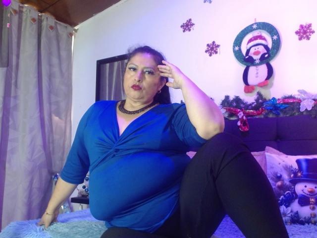 Keyla69Hot, fat chick with blue top, black pants, and necklace, sits on bed, hand on head, looking at camera, Christmas decor, penguin wall art, purple bed pillow.