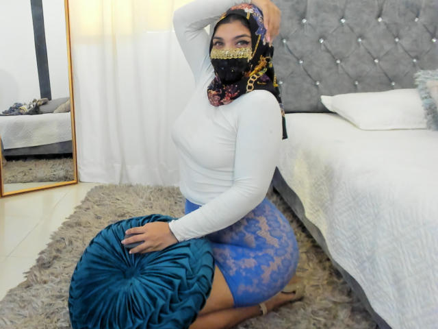Aniisa Ali mit dunklem Haar, die eine schwarze Maske mit goldener Verzierung, ein weißes Oberteil mit langen Ärmeln und einen blauen Spitzenrock trägt, kniet auf einem Teppich, hält ein türkisfarbenes Kissen und befindet sich neben einem Bett..