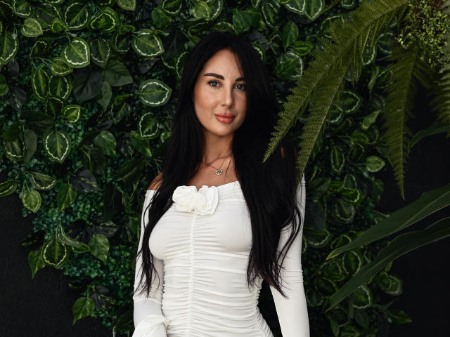 KimberlyWorld has long black hair, white off-shoulder dress with rose, small tits, and a necklace, standing in front of green foliage. She's smiling with a cute, sexy look.