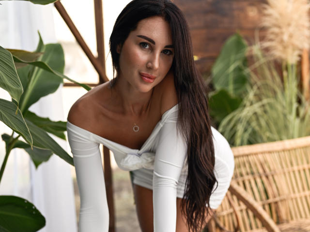 KimberlyWorld with long dark hair, white off-shoulder top, and white shorts, bending forward, showing her small ass and cute face, smiling softly, green plant in background.