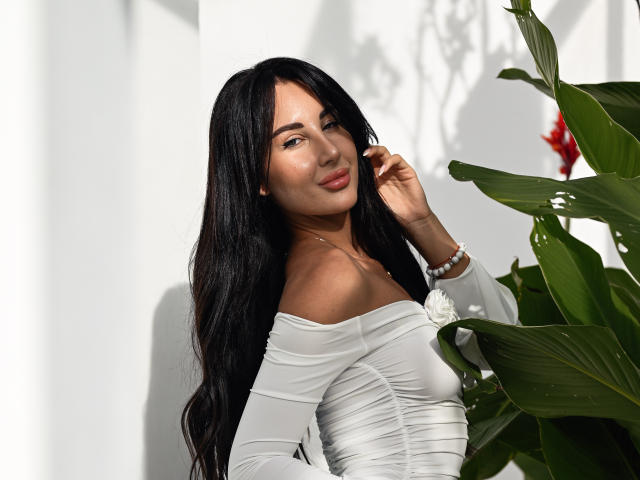 KimberlyWorld with long black hair, white off-shoulder dress, small white flower, bracelet, smiling, touching her hair, green plant with red flower on the right.