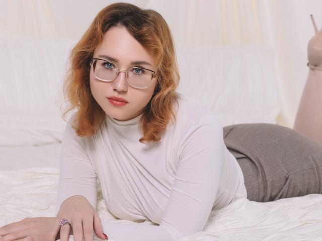 TeresaBirt with curly red hair, glasses, white turtleneck, gray shorts, lying on a bed, looking at camera, hands on bed, pink ring, white sheets, relaxed, cute.