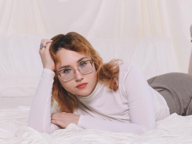 TeresaBirt with glasses, lying on white bed, wearing a white turtleneck and gray pants, hand on head, looking at camera, red lips, wavy orange-brown hair, soft light.