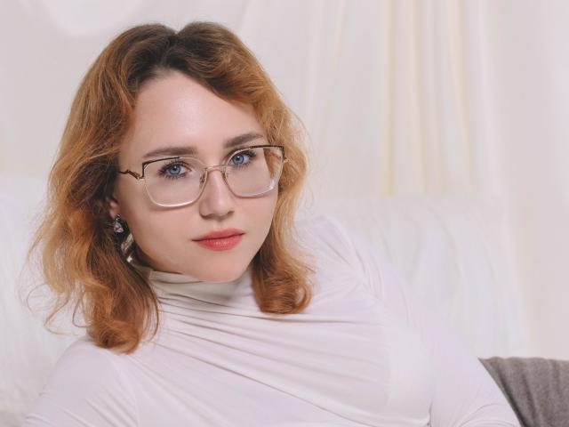 TeresaBirt with wavy red hair, glasses, white turtleneck, and red lipstick, sitting on a white couch. Her blue eyes and earrings are visible.
