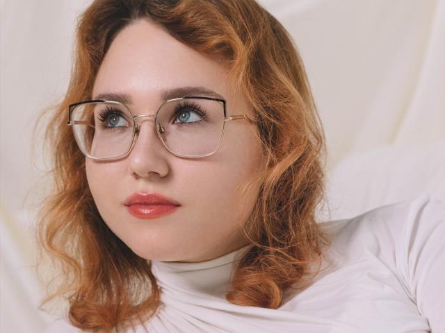 TeresaBirt with red curly hair, wearing glasses and a white turtleneck, has blue eyes and red lips, looking slightly to the side with a calm expression.