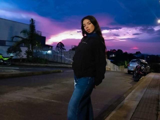 KatherinJohns with long brown hair, black hoodie, and blue jeans stands on a street at dusk, looking down with a slight pout, hands in pockets, next to parked bikes.