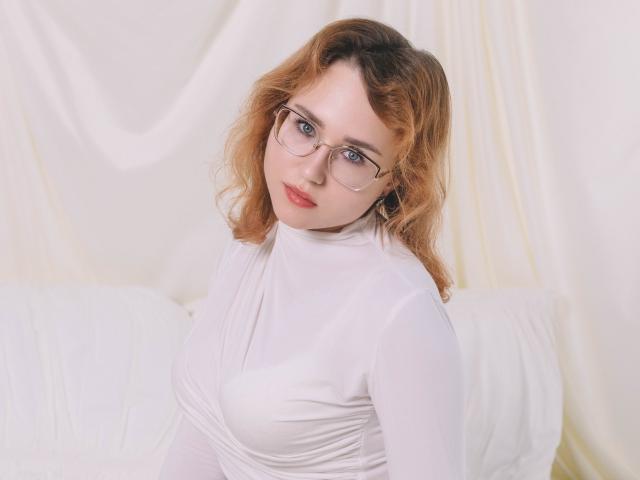 TeresaBirt with curly red hair, glasses, and white dress, showing her small tits and looking up with a slight smile. Soft, white background.