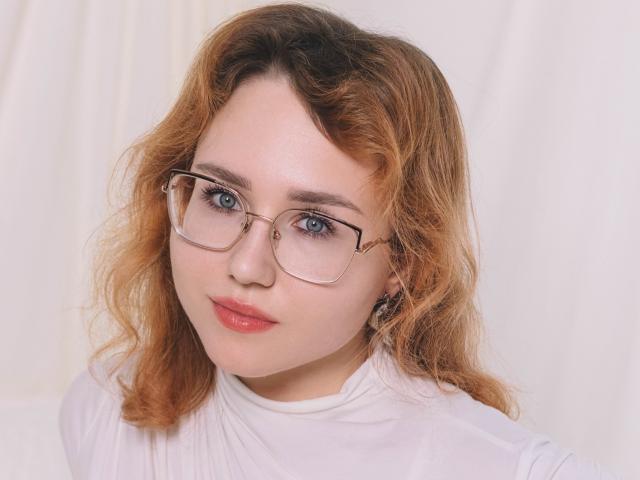 TeresaBirt has wavy orange hair, wears glasses with a gold frame, and has blue eyes, pink lips, and a white shirt. Her ass is not visible.