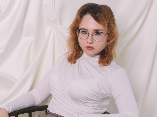 TeresaBirt with red hair, glasses, and white turtleneck, showing her small tits, sitting with a serious look, hands on chair.