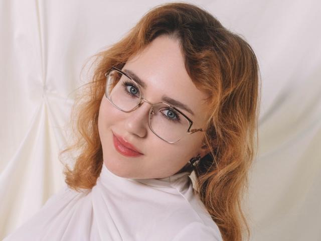 TeresaBirt with wavy red hair, glasses, and white turtleneck, smiling with pink lips, blue eyes, and fair skin. Her tits are not visible, but she's cute.