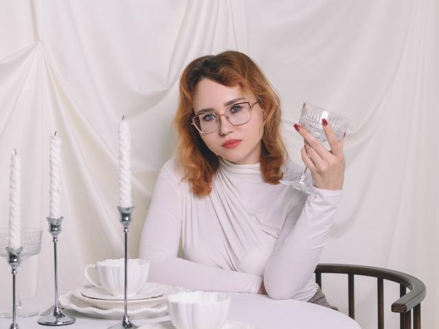TeresaBirt with glasses, red nails, white dress, holding a glass, sitting at a table with candles, cups, and plates, looking at the camera.