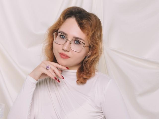 Teresa Birt, com cabelo laranja ondulado, óculos, blusa branca, unhas vermelhas e um anel roxo, sorrindo suavemente, com a mão perto do queixo. Seus seios são pequenos e sua vulva não está visível..