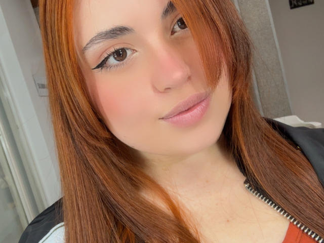VicluX has orange hair, brown eyes with black eyeliner, pink lips, and a slight smile. She's wearing a black and white jacket with a zipper.