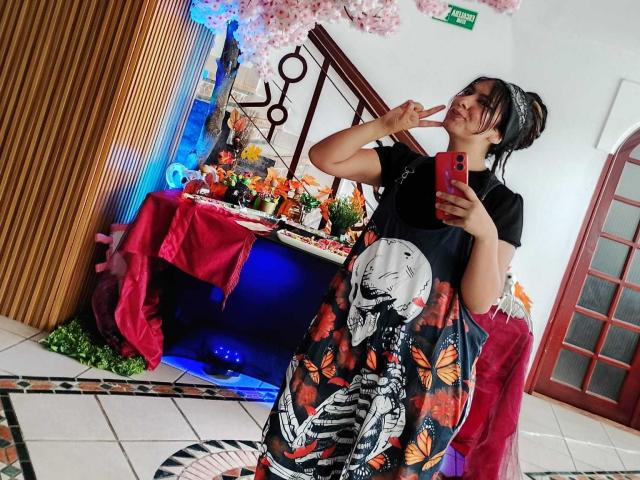 BeckyHuston with black top, floral and skull-patterned skirt, holding red phone, making peace sign, standing in front of altar with pink cherry blossom decor.