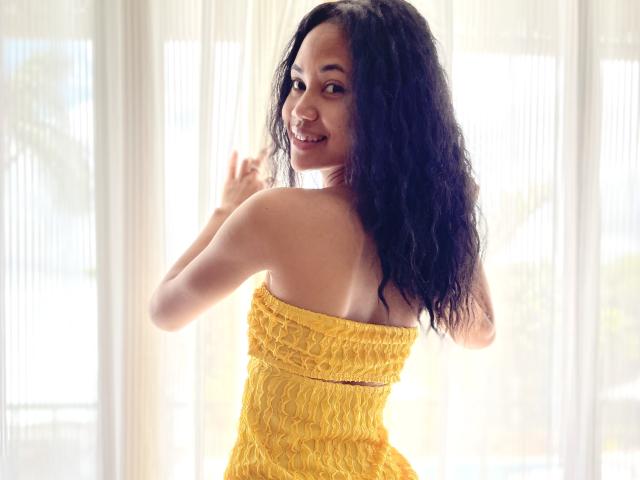 AubreyLoo with wavy black hair, smiling at the camera, wearing a yellow strapless dress, showing her bare back and yellow dress, standing in front of sheer curtains.