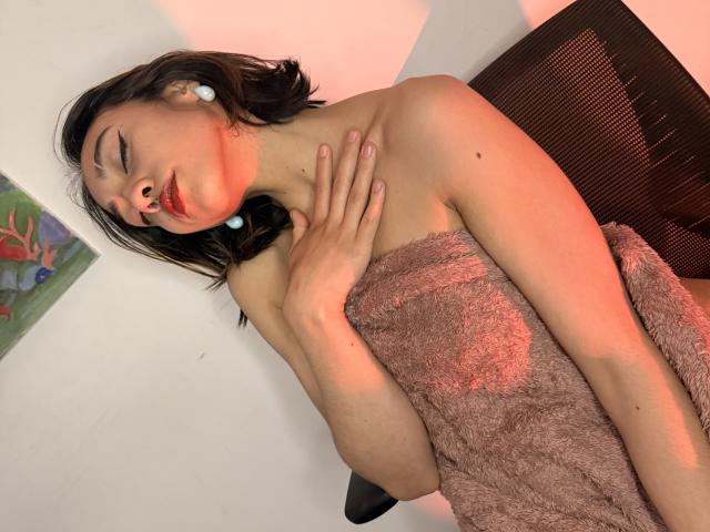 CamiVargas with wet brown hair, red lipstick, and blue earbuds, wrapped in a pink towel, hand on neck, sitting in a chair with red lighting on her skin.
