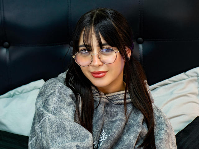 SopphieBloom with long black hair, glasses, and red lipstick, wearing a gray hoodie, sitting on a bed, smiling softly. Her tits are small and her pussy is not visible.