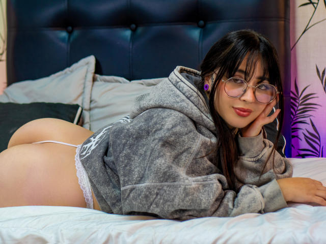 SopphieBloom with glasses, lying on a bed in a grey hoodie, white lace thong, and purple earrings, showing her ass, smiling, and resting her head on her hand.