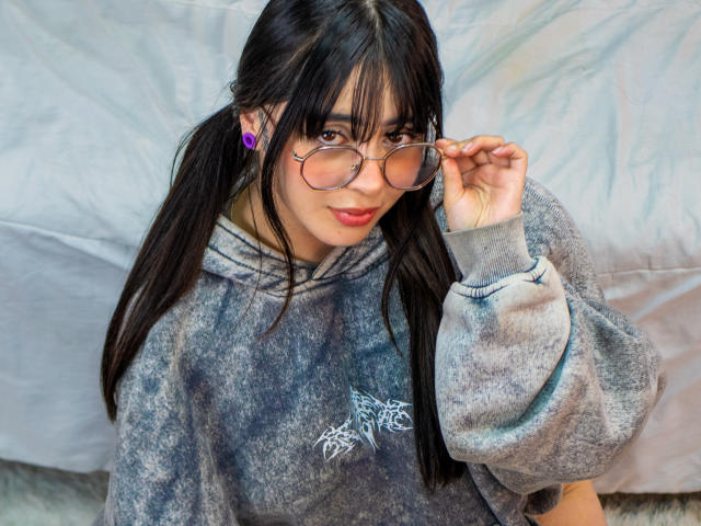 SopphieBloom with pigtails, wearing a gray hoodie, holding glasses, purple earbuds, and a cute smile, showing her tits, with a fluffy gray background.