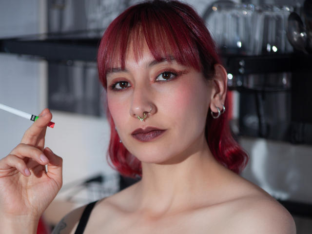 AliRousse, con cabello rojo, un piercing en la nariz y aretes con forma de corazón, sostiene un cigarrillo encendido, tiene lápiz labial rosa y está mirando a la cámara con una ligera sonrisa..