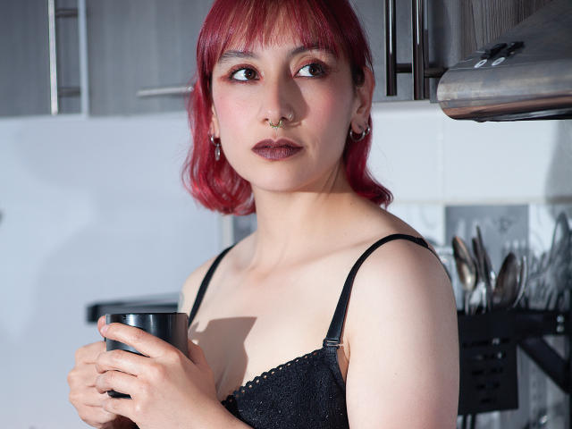 AliRousse, avec des cheveux roux, un anneau au nez et du rouge à lèvres foncé, tenant une tasse, portant un soutien-gorge en dentelle noire, regardant sur le côté, dans une cuisine. Seins visibles, de taille moyenne..