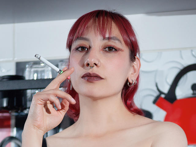AliRousse with red hair, nose ring, and ear piercings, holding a lit cigarette to her right nostril, has dark lipstick and a confident expression.