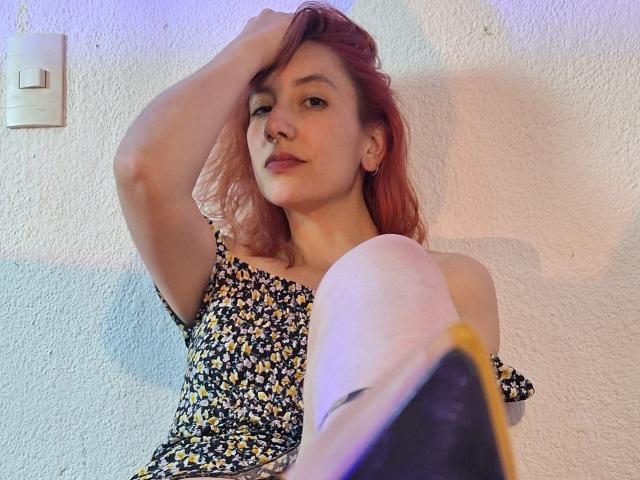 AliRousse, con i capelli rosa, indossa un abito floreale, ha una mano tra i capelli e una gamba in primo piano, mostrando una parete bianca e un interruttore della luce..