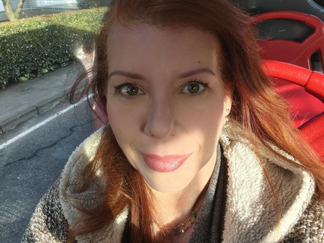 Khharha has red hair, green eyes, and is wearing a fluffy beige coat with a brown shirt. Her pink lips are slightly upturned in a smile. She's in a car.